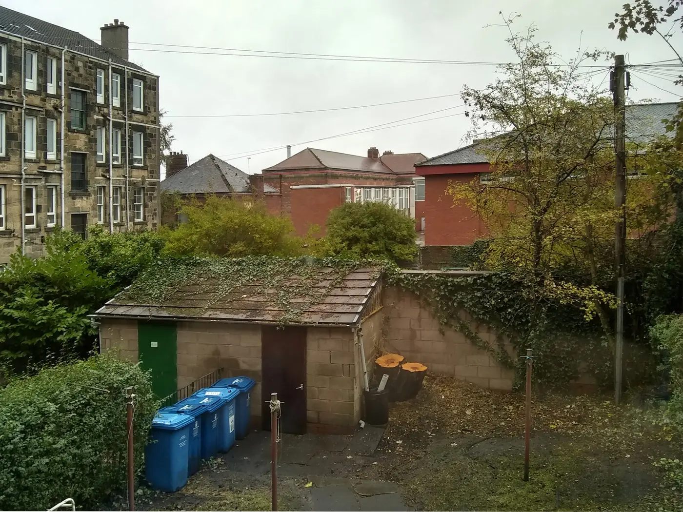 The back court behind Glasgow sandstone tenements, with a communal bin store, drying poles and the rear gable of the tenement beyond.